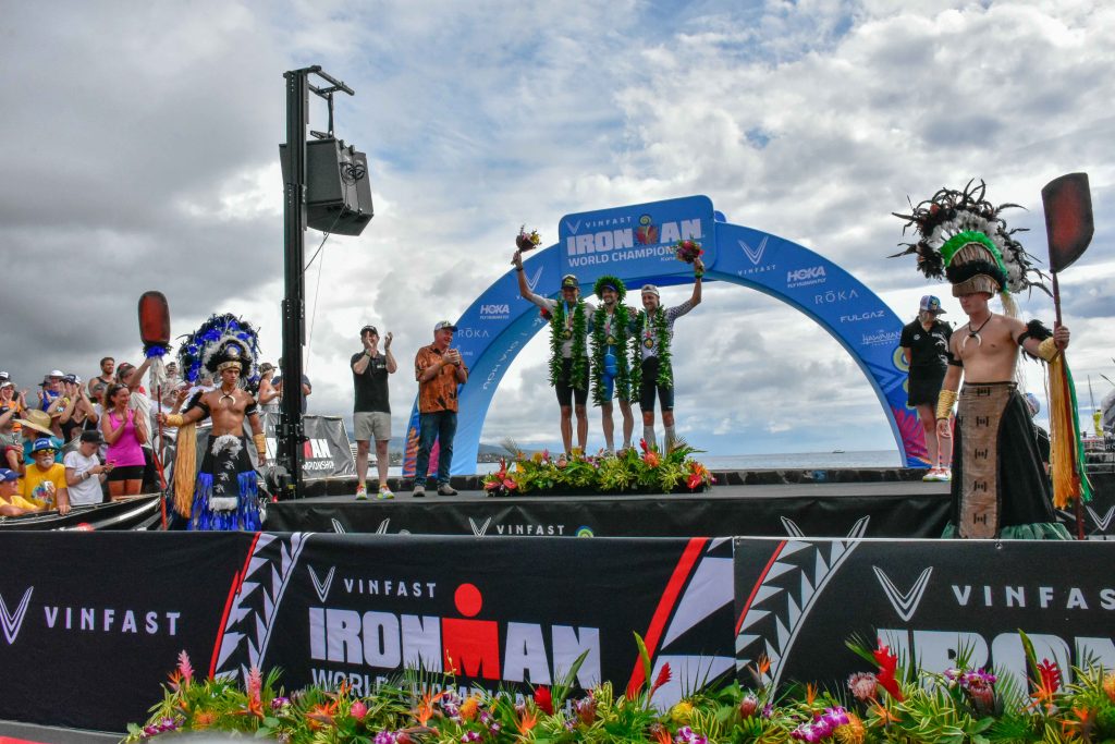 The 2024 Ironman World Championship winner Patrick Lange (hands raised) is on the podium with second-place Magnus Ditlev of Denmark (left) and third-place Rudy von Berg of the United States as Hawai'i County Mayor Mitch Roth and Ironman CEO Scott DeRue look on. (Cammy Clark/Big Island Now)