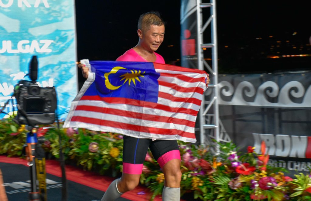 Toi "Gary" Wong finished the 2024 VinFast Ironman World Championship in his hometown of Kona in a time of 12:23:09, but across the finish line he carried the flag of his home country, Malaysia. (Cammy Clark/Big Island Now)